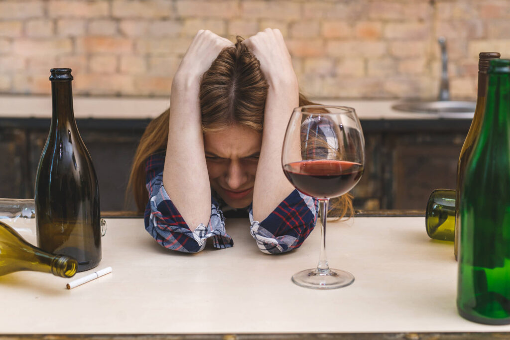 Zrozpaczona kobieta wśród butelek z głową w dłoniach - objawy alkoholizmu