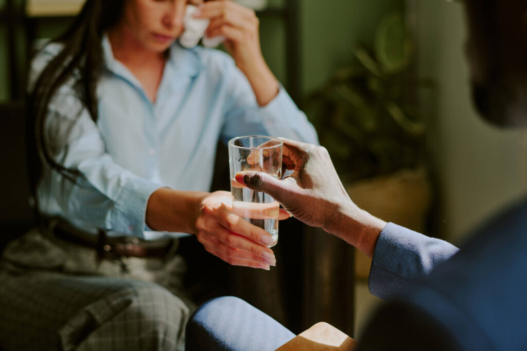 Dłoń mężczyzny podająca szklankę wody zapłakanej kobiecie - wsparcie bliskich po leczeniu alkoholizmu
