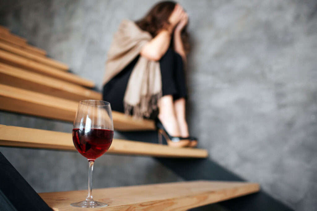 Kieliszek wina na pierwszym planie i kobieta siedząca na schodach z głową w dłoniach - picie alkoholu jako ucieczka od depresji