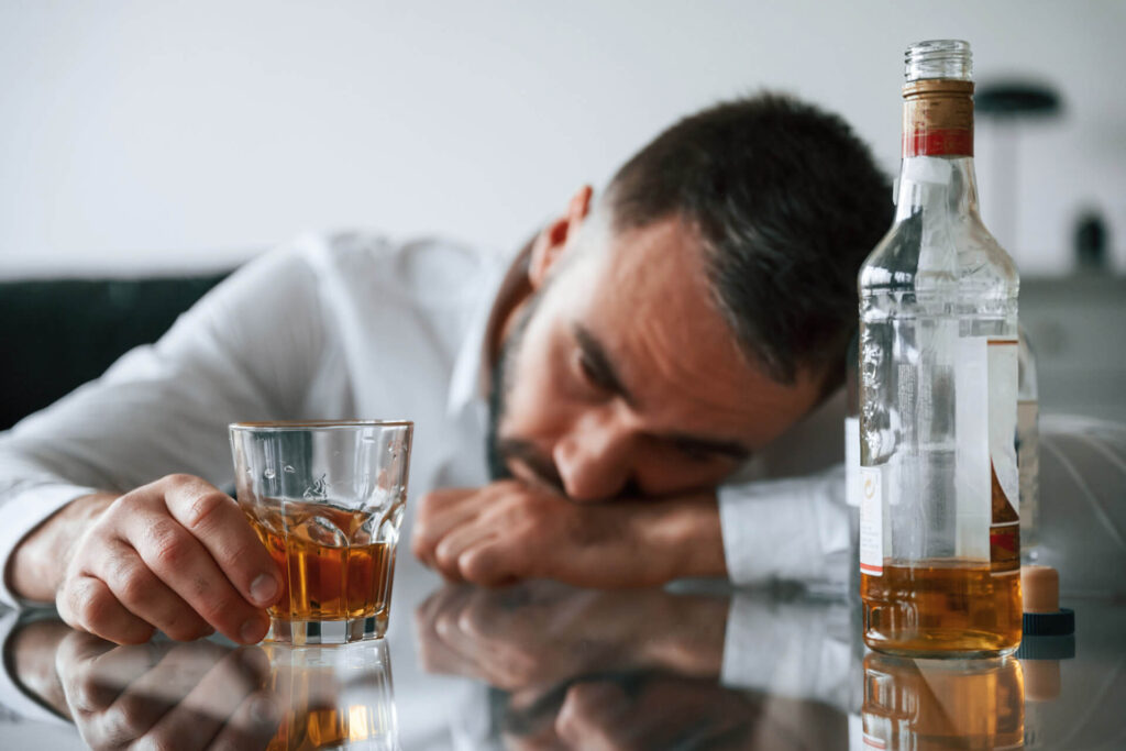 Mężczyzna leżący na barze, wpatrzony w szklankę, z butelką alkoholu na pierwszym planie - objawy uzależnienia od alkoholu