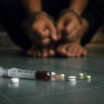 Substancje psychoaktywne leżące na podłodze, w tle zaciśnięte dłonie osoby uzależnionej od narkotyków